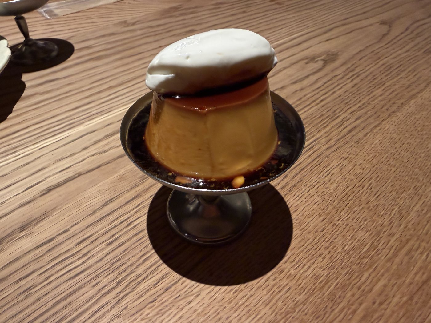 木のテーブルに置かれた、銀色の器に乗ったプリン。上に生クリーム