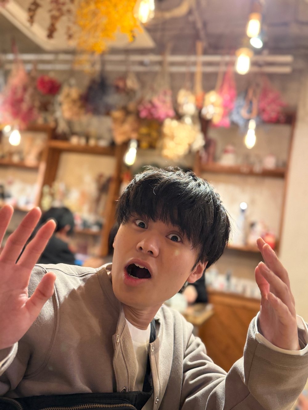 ドライフラワーが吊るされた店内で、筆者が目を見開き、口を開けて両手を上げている。背景に他のお客さんが2人写っている